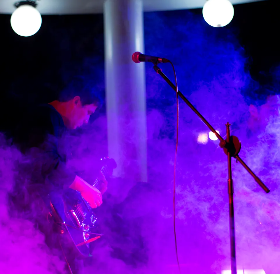 Músico tocando guitarra entre humo e iluminación violeta durante un concierto.