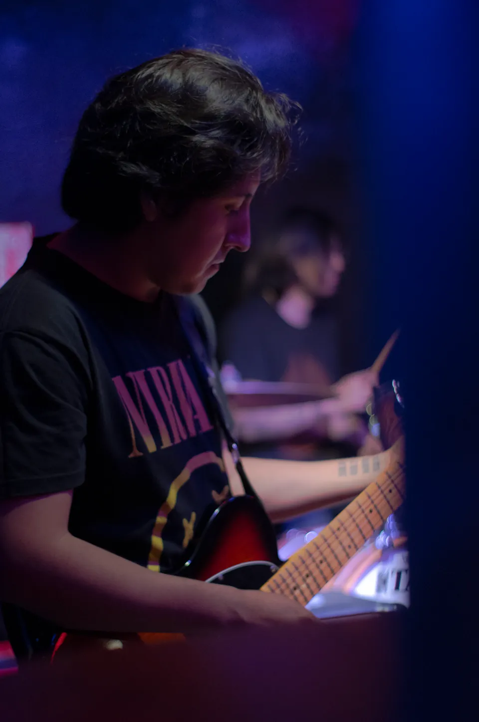 Guitarrista concentrado en su instrumento con un baterista al fondo.